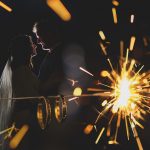 una pareja de novios agarrados de la cintura, mirándose a los ojos se ven al fondo y en primer plano están sus anillos iluminados por las chispas de una bengala