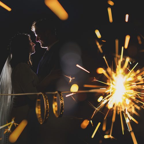 una pareja de novios agarrados de la cintura, mirándose a los ojos se ven al fondo y en primer plano están sus anillos iluminados por las chispas de una bengala